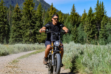 A black male rider wearing sunglasses and a cap rides an Aniioki A8 60V electric bike with a fat tire on a rugged dirt trail.