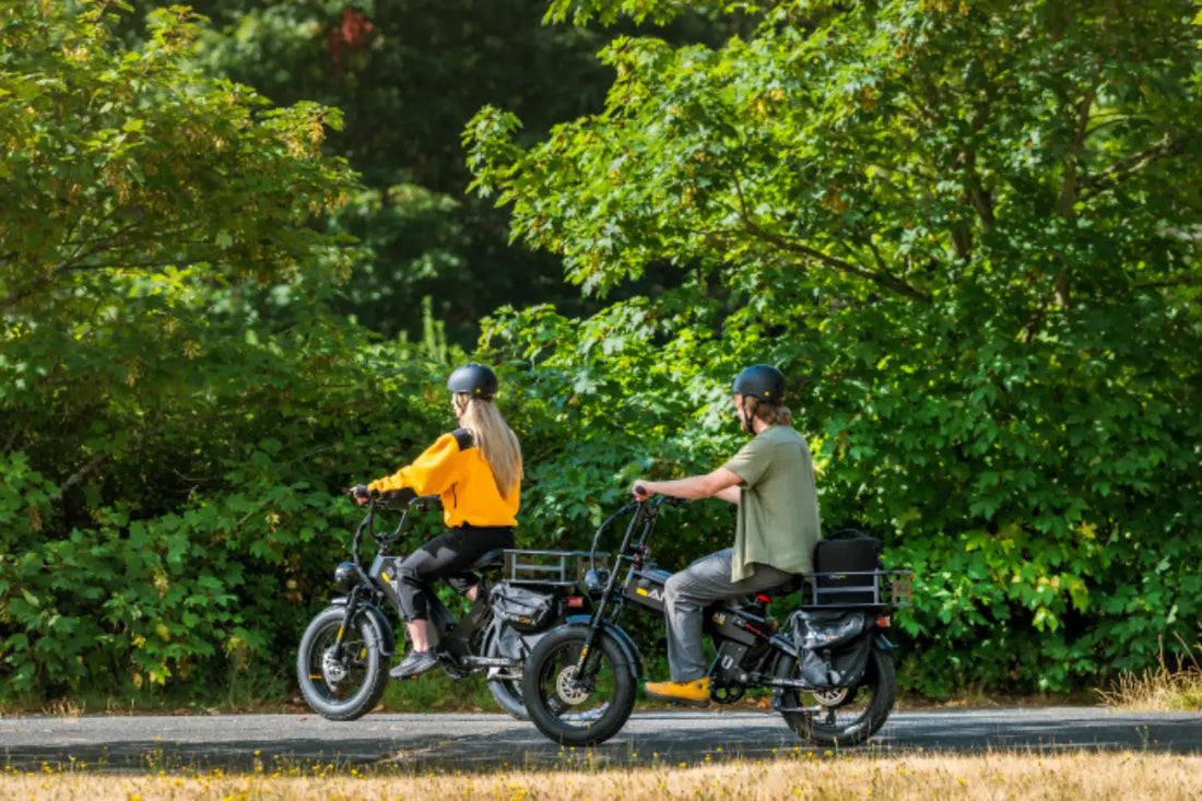 A Caucasian man and woman riding black Aniioki AQ177 48V long range electric bikes on a paved park trail surrounded by green trees
