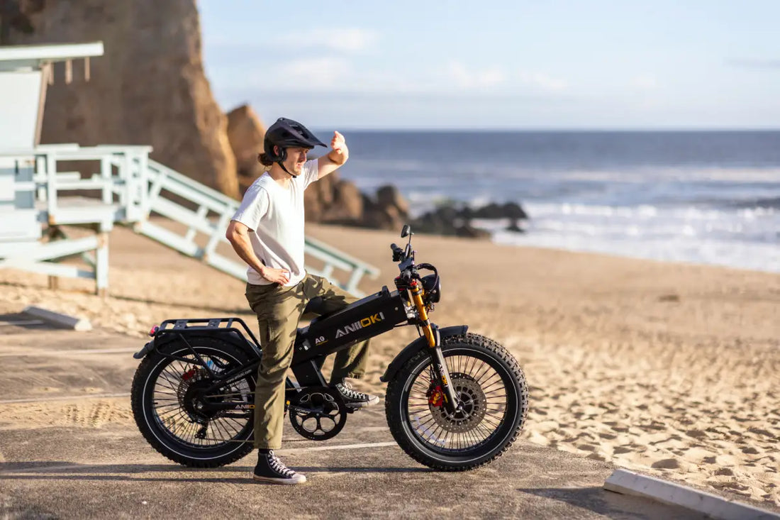  Aniioki A9 black electric bike with high-performance E-bike Sine Wave Controller, rider in white shirt and green pants standing on a coastal beach path with ocean waves and lifeguard tower backdrop