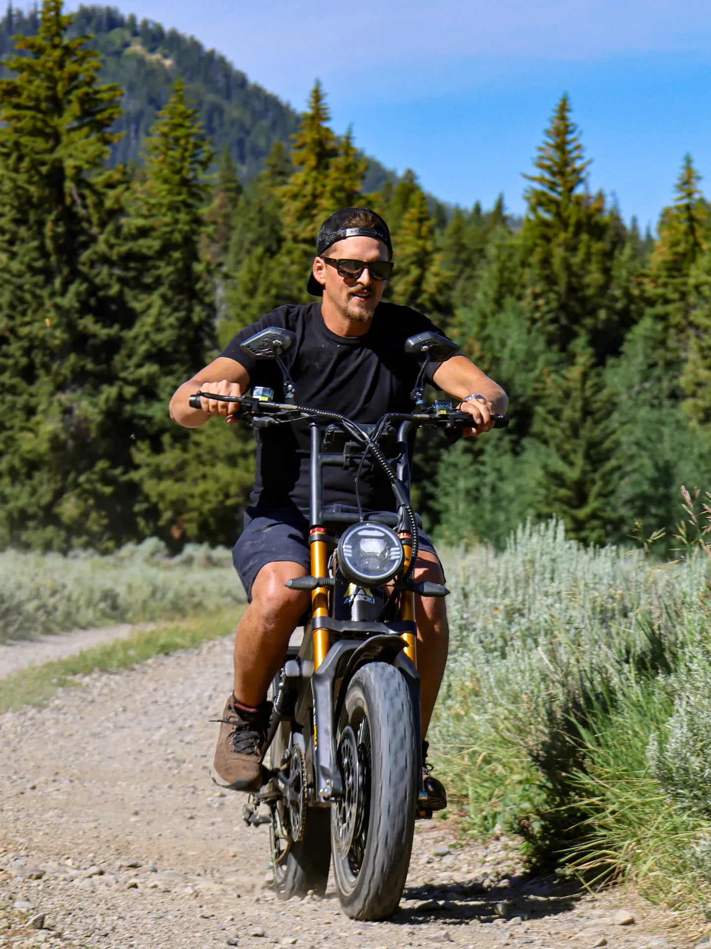 A man riding an electric bike on a gravel path surrounded by green trees and brush under a blue sky.