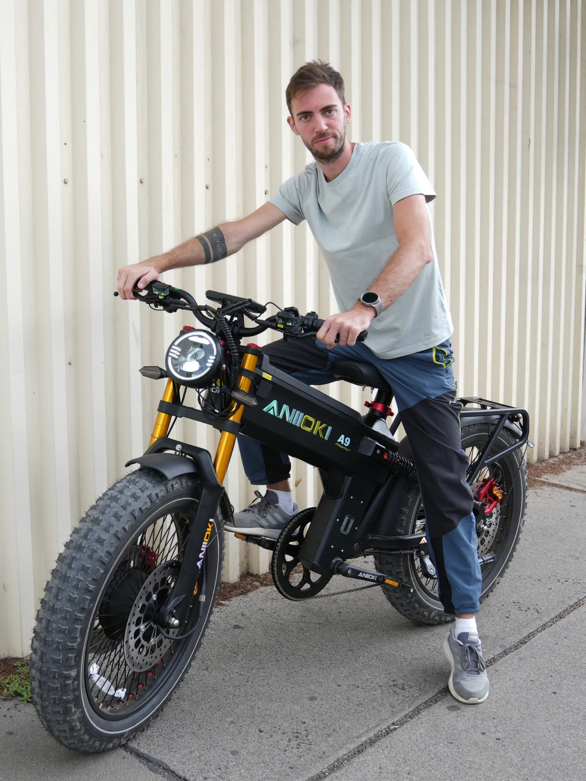 A man wearing a light gray t-shirt and dark pants sits on a black Aniioki eBike with knobby tires, positioned in front of a corrugated metal wall.